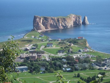 Village of Percé and Percé Rock seen from the top of a mountain. There are a lot of lawns all around the village.